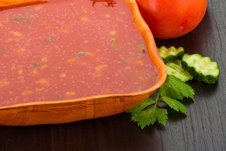 A vibrant bowl of gazpacho soup, garnished with cucumber slices and fresh parsley, sits on a dark wooden surface alongside a tomato.の写真素材