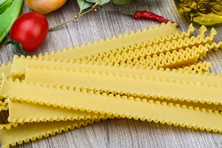 A pile of uncooked ruffled edge lasagna noodles rests on a wooden surface, surrounded by fresh vegetables and spices.の写真素材