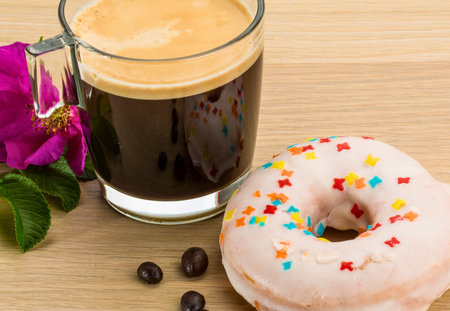 A close-up shot of a glass of dark coffee with foam, a frosted donut with sprinkles, a flower, and coffee beans on a wooden surface.の写真素材
