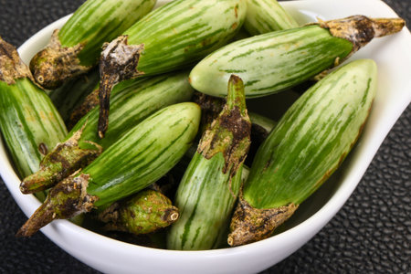 A close-up shot of numerous small, vibrant green baby eggplants with striped skin, piled in a white ceramic bowl.の写真素材