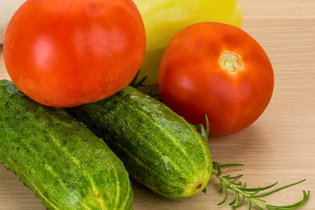 A close-up shot of vibrant red tomatoes, crisp green cucumbers, and a sprig of fresh rosemary arranged on a light wooden background.の写真素材