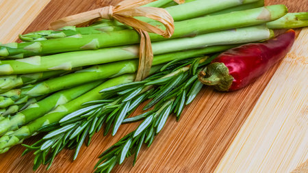 A bundle of fresh green asparagus spears is tied with twine and adorned with rosemary, alongside a vibrant red chili pepper on a wooden surface.の写真素材