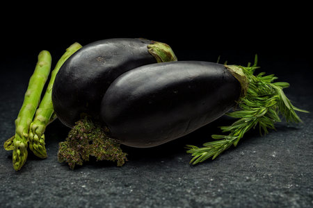 Two dark eggplants are presented with vibrant green asparagus spears and fresh herbs on a textured dark background.の写真素材