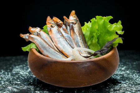 A rustic wooden bowl overflowing with small, silver fish and a vibrant green lettuce leaf, presented against a dark, textured background.の写真素材