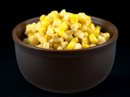A dark brown ceramic bowl filled with bright yellow, cooked corn kernels against a solid black background.の写真素材