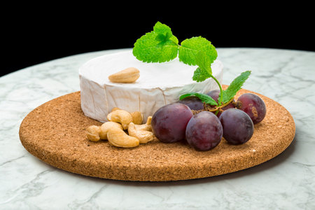 A round of creamy brie cheese is artfully arranged with fresh grapes and crunchy cashews on a cork serving board.の写真素材
