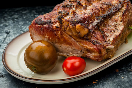 A close-up shot of a succulent, roasted pork shoulder served with a bright red cherry tomato and a dark olive on a platter.の写真素材