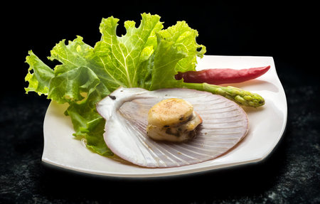 A single scallop sits in its shell, garnished with crisp lettuce, asparagus, and a red chili pepper on a white plate.の写真素材