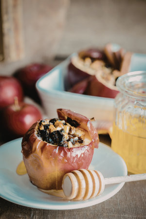 A close-up of a baked apple topped with nuts and dried fruits, drizzled with honey, placed on a white plate with a honey dipper. Surrounded by fresh apples and additional baked apples in the background.の写真素材