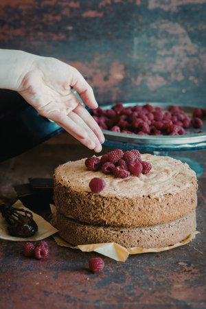 A close-up image of a hand placing fresh raspberries on top of a two-layer cake. The cake is being decorated with raspberries and chocolate pieces, and a bowl of additional raspberries is visible in the background.の写真素材