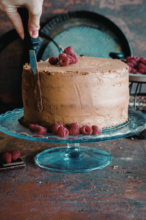 A hand is cutting a rich chocolate cake decorated with fresh raspberries on a blue cake stand. The...