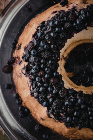 A close-up image of a beautifully baked bundt cake topped with fresh blueberries and blackberries. The cake has a golden-brown crust and is presented on a dark plate, with a few berries scattered around it.の写真素材