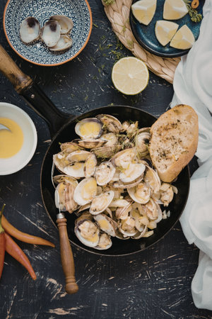 A beautifully arranged clam dish served with lemon slices, bread, and various seasonings. The clams are presented in a black bowl, accompanied by a side of bread, lime, chili peppers, and a small bowl of oil.の写真素材