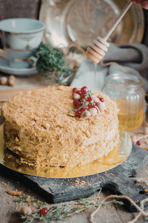 A beautifully crafted honey cake sits on a wooden board, adorned with fresh berries and a honeycomb decoration. Honey is being drizzled over the cake, adding a sweet touch. Surrounding the cake are various kitchen items, including a teacup, jars of honey, and herbs.の写真素材