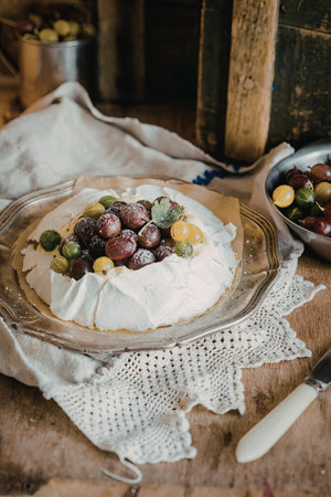 A beautifully presented pavlova topped with whipped cream and an assortment of fresh berries, including grapes and cherries. The dessert is placed on a rustic wooden table with a lace doily and a knife nearby.の写真素材