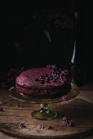 A beautifully crafted purple cake sits elegantly on a glass stand, surrounded by delicate purple flowers and a dark, moody background.の写真素材