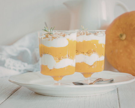 A visually appealing pumpkin parfait served in elegant glass cups, garnished with a sprig of rosemary, placed on a white plate with a pumpkin in the background.の写真素材