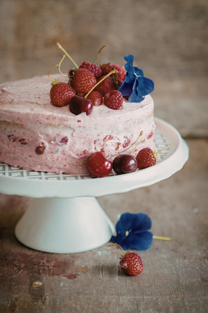 A beautifully decorated berry cake with pink frosting sits on a white pedestal. The cake is adorned with fresh strawberries, raspberries, cherries, and a single purple flower.の写真素材