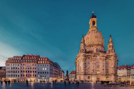 A stunning image of the Dresden Cathedral, also known as the Frauenkirche, standing majestically in a bustling city square. The cathedral's intricate architecture is highlighted against a deep blue sky, with surrounding buildings and people adding to the lively atmosphere.の写真素材