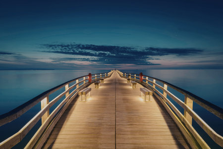 A serene evening scene featuring a long wooden pier illuminated by soft lights, extending into a calm body of water under a deep blue sky.の写真素材