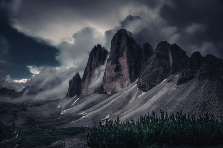 A breathtaking view of a rugged mountain range partially shrouded in mist and clouds, with dramatic...
