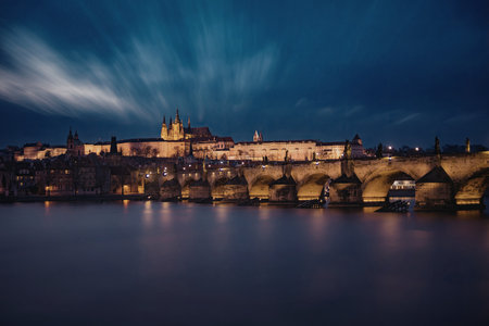 A serene evening view of Prague Castle and Charles Bridge, illuminated against a deep blue sky with scattered clouds reflecting on the calm river below.の写真素材