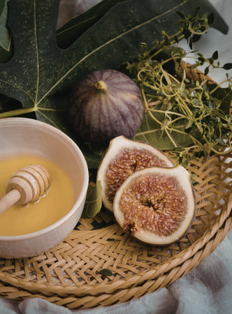 A close-up image of fresh figs, one sliced open, accompanied by a bowl of honey and a honey dipper on a woven tray, surrounded by green leaves.の写真素材