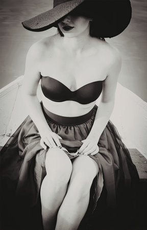 A black and white photograph of a woman wearing a vintage-style outfit, including a wide-brimmed hat, strapless top, and long skirt. She is seated with her legs crossed, exuding an air of sophistication and grace.の写真素材