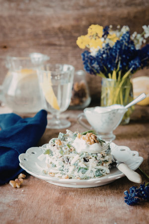A plate of fresh salad with walnuts and a creamy dressing, accompanied by a lemon slice, a blue napkin, and a vase of colorful flowers on a rustic wooden table.の写真素材