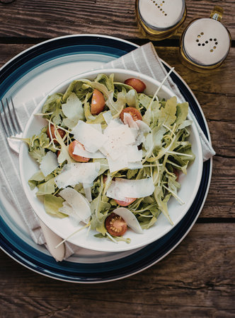 A vibrant salad featuring fresh greens, ripe tomatoes, and shavings of cheese, served on a white plate with a fork. The plate is accompanied by small containers of salt and pepper.の写真素材