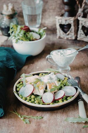 A vibrant salad featuring peas and radishes, served in a bowl on a rustic wooden table. Accompanied by a glass of water, a small dish of dressing, and a fork.の写真素材
