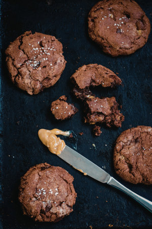 A close-up image of freshly baked chocolate cookies sprinkled with sea salt, one of which is partially eaten, lying on a dark surface with a knife and some spread nearby.の写真素材