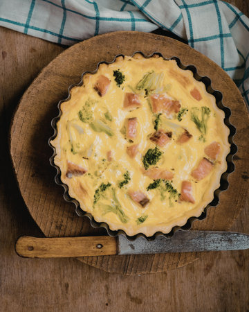 A close-up image of a freshly baked vegetable quiche featuring broccoli, tomatoes, and cheese, placed on a wooden board with a knife beside it.の写真素材