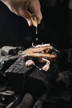 A hand squeezing lemon juice over a piece of grilled meat placed on charred wooden planks,...
