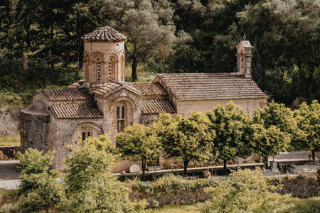 An old stone church with a dome and bell tower stands amidst dense, verdant trees and vegetation,...