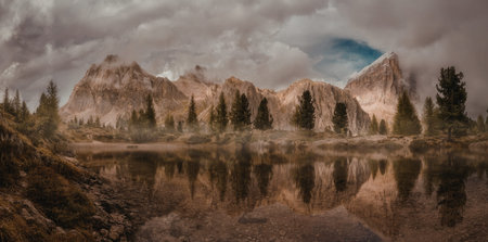 A serene mountain landscape featuring towering peaks under a cloudy sky, with a calm pond reflecting...