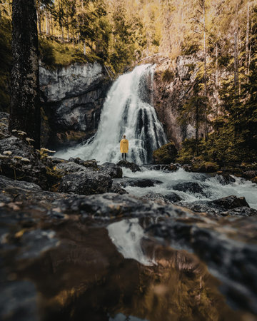 A person in a yellow jacket stands near a waterfall surrounded by dense forest and rocky terrain.