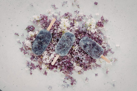 A close-up image of three purple popsicles placed on a bed of flower petals, creating a visually...