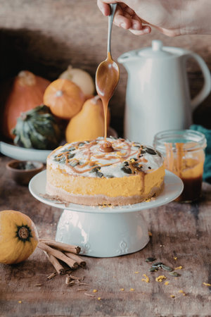 A delicious pumpkin cheesecake is being drizzled with caramel sauce. The cake is topped with whipped cream and pumpkin seeds, and is surrounded by pumpkins, cinnamon sticks, and a teapot.の写真素材
