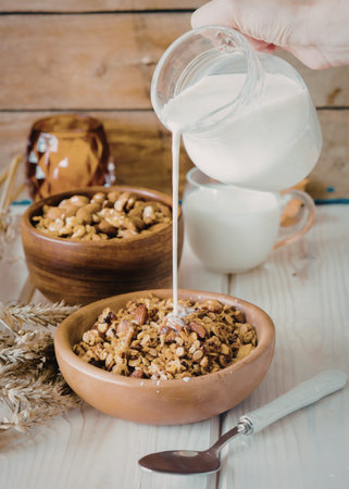 A hand is pouring milk from a glass pitcher into a bowl filled with granola. The bowl is placed on a...