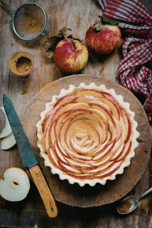 A beautifully arranged apple tart sits on a wooden table surrounded by whole apples, a knife, and...