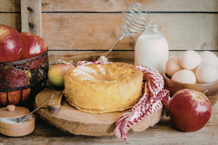 A freshly baked apple cake sits on a wooden table surrounded by fresh apples, eggs, milk, and a...