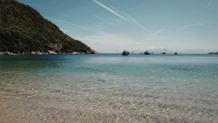 A picturesque coastal scene featuring crystal-clear turquoise waters, a sandy beach, and a lush green hillside under a bright blue sky.の写真素材