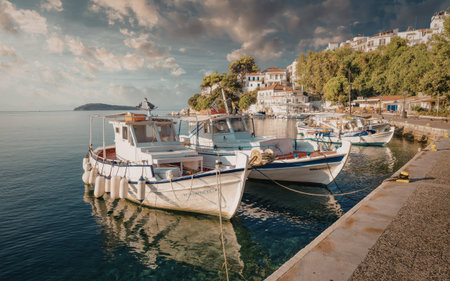 A tranquil harbor scene featuring several small boats moored at a dock. The calm water reflects the boats and the surrounding buildings. The sky is partly cloudy, with a mix of clouds and blue sky, indicating a peaceful day.の写真素材
