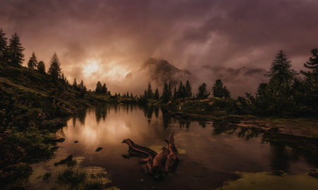 A tranquil mountain lake reflects the warm hues of a setting sun, surrounded by dense forests and...