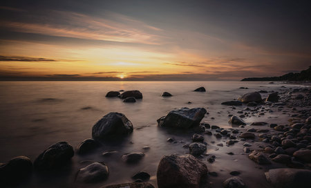 A tranquil beach scene at sunset with rocks scattered along the shore and gentle waves lapping...