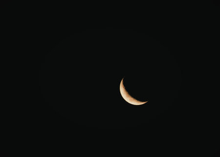 A thin crescent moon is visible against a dark night sky. The moon is illuminated on the right side,...