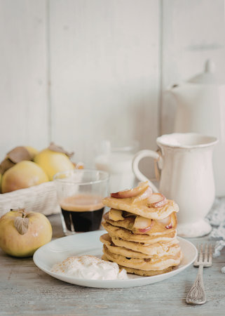 A stack of golden-brown apple pancakes topped with fresh apple slices, served on a white plate with a dollop of cream and a sprinkle of cinnamon. Surrounding the plate are fresh apples in a basket and a white pitcher.の写真素材