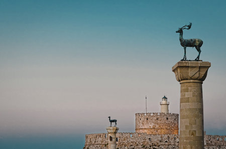 A stone structure featuring a prominent deer statue stands against a backdrop of a serene sunset. Additional smaller statues are visible on the structure, with a lighthouse in the distance.の写真素材