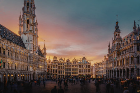 A vibrant sunset illuminates the historic Grand Place in Brussels, showcasing its ornate guild houses and the majestic Town Hall. The square is bustling with people enjoying the picturesque evening.の写真素材
